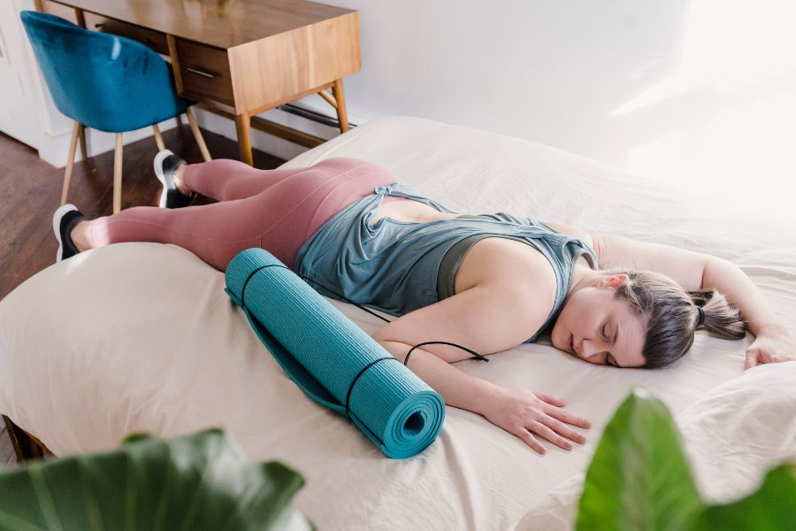 Woman in workout clothes laying face down on her bed with her yoga mat